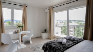 Un dormitorio moderno con una cama grande, una pequeña mesa redonda con sillas, una mesita de noche y vistas de la ciudad a través de amplias ventanas.