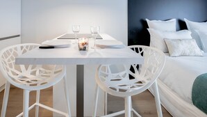 Una habitación moderna con una mesa blanca dispuesta para dos personas, dos sillas blancas, una cama con ropa de cama blanca y una pared de acento azul oscuro.
