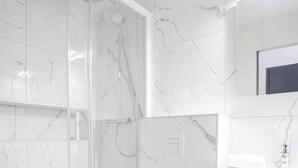 Un baño moderno con cabina de ducha de vidrio, azulejos de mármol blanco, inodoro blanco y tocador minimalista con espejo.