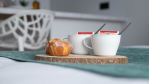Un desayuno con un muffin y dos tazas blancas sobre un posavasos de madera, una silla blanca y un paño verde al fondo.