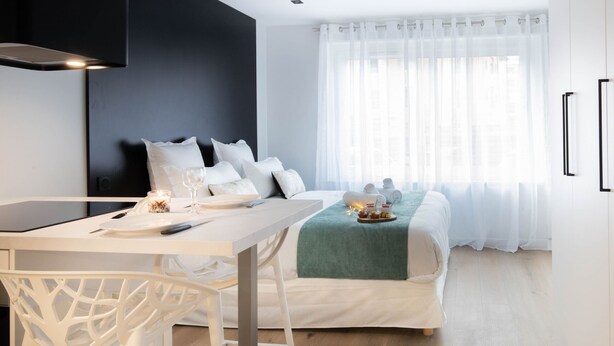 Una habitación de hotel moderna con comedor, una cama con una manta verde y un amplio ventanal con cortinas translúcidas.