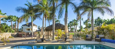 A pool area with lounge chairs and umbrellas, surrounded by palm trees and a building with a thatched roof.