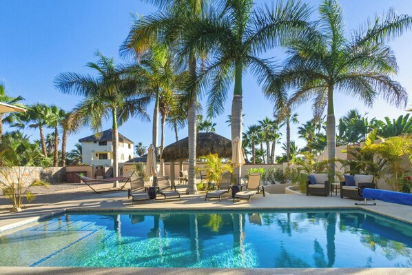 A pool area with lounge chairs and umbrellas, surrounded by palm trees and a building with a thatched roof.