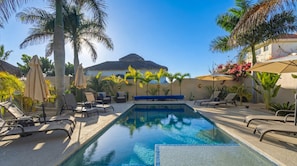 A pool area with lounge chairs and umbrellas, surrounded by palm trees and a building with a thatched roof.