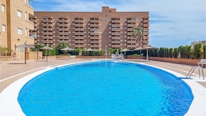 Una piscina amplia y cristalina rodeada de sillas de descanso y sombrillas, con un edificio de varios pisos al fondo.