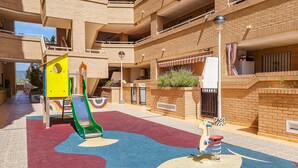 Zona de juegos infantiles con un tobogán y un caballito balancín, rodeada por un edificio de varios pisos con balcones y fachada de ladrillo.