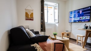 A living room with a black sofa, a patterned throw, a small round table with a plant, and a television mounted on the wall. - Secrets d ambre rue royale (Annecy)
