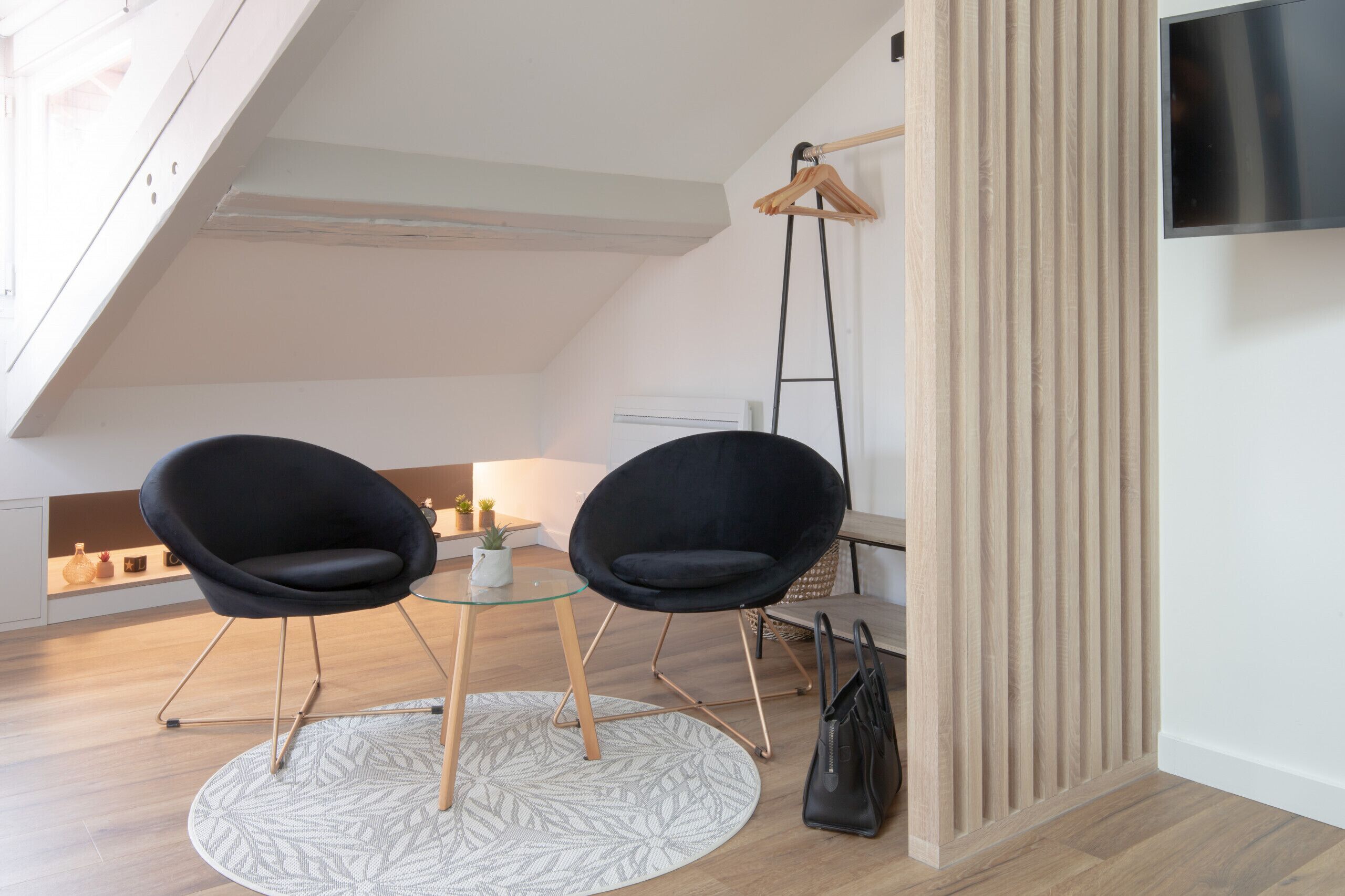 Een moderne loft slaapkamer met schuine plafonds, een glazen tafel met twee zwarte stoelen, een televisie aan de muur en een houten vloer.