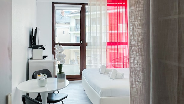 Une chambre d’hôtel moderne avec un lit blanc, une petite table ronde ornée d’un vase de fleurs et une vue sur la ville, visible à travers une grande fenêtre.