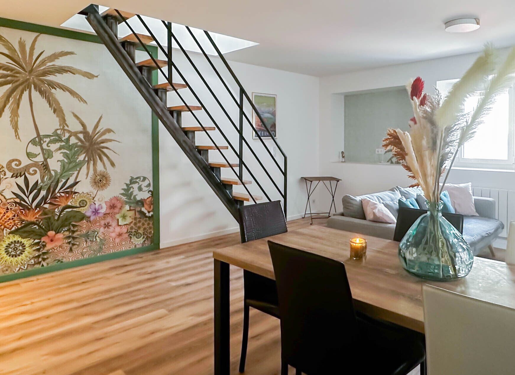 Een moderne leefruimte met een houten vloer, een trap met zwarte leuningen, een eethoek met tafel en stoelen, een bank en een decoratieve wand met tropische planten.