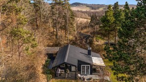 En hytte med skråt tag, omgivet af tæt skov og bakker i baggrunden.