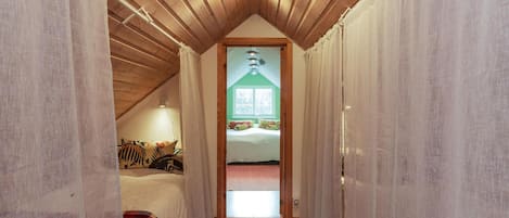A narrow hallway with wooden ceiling beams, a bed with colorful bedding, and a window with a view of trees.