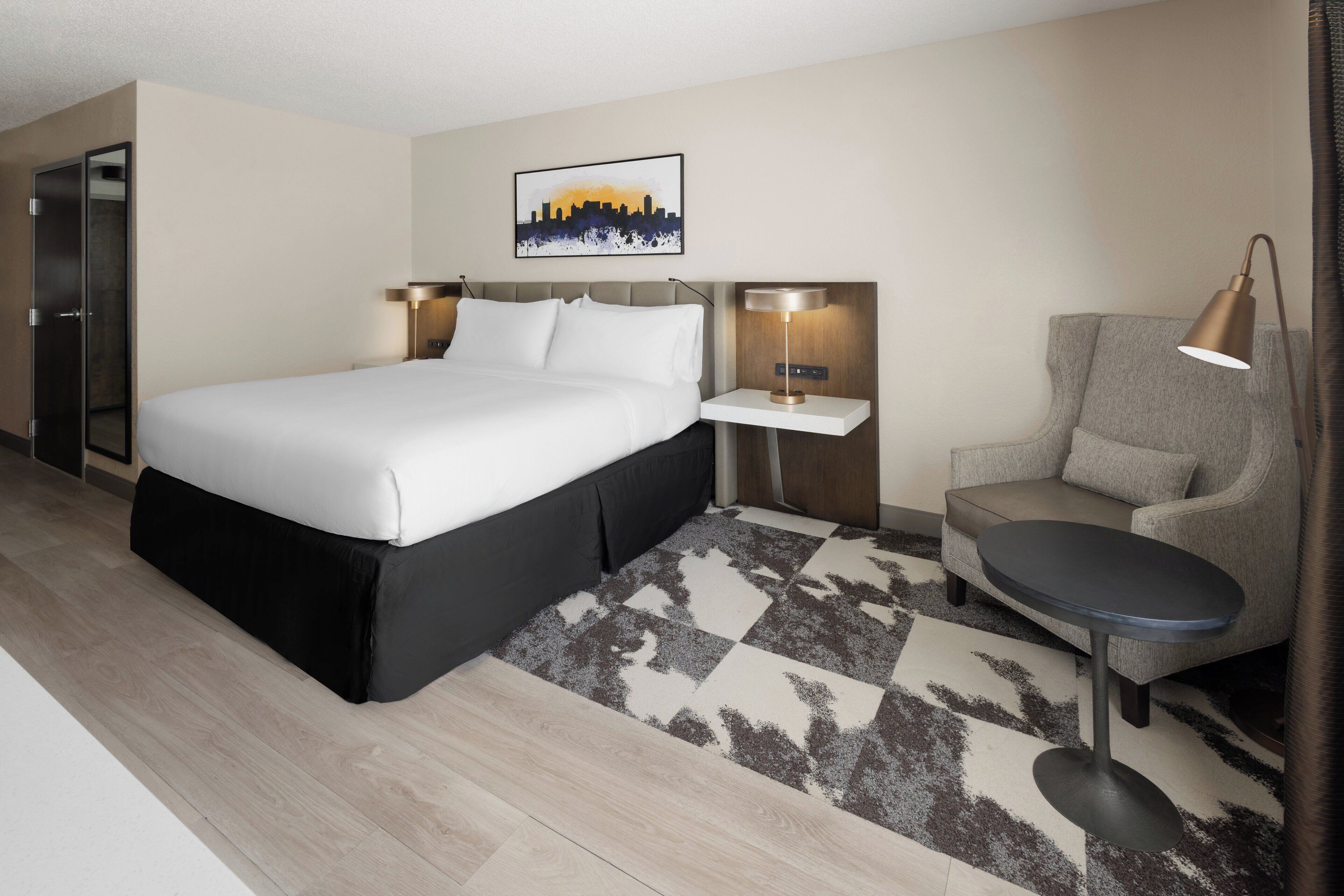 A modern hotel room with a large bed, a small table, a chair, and a cityscape artwork on the wall.