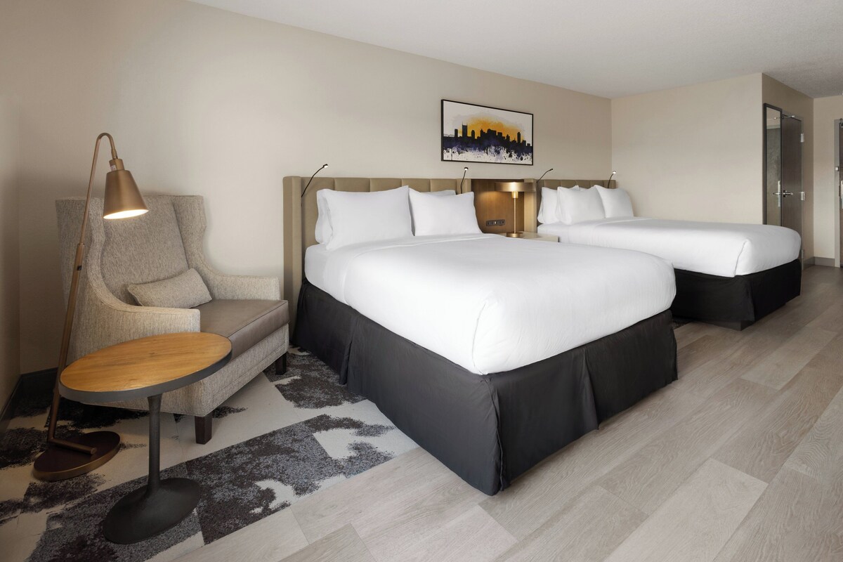 A hotel room with two beds, a sofa, a desk, and a cityscape artwork on the wall.