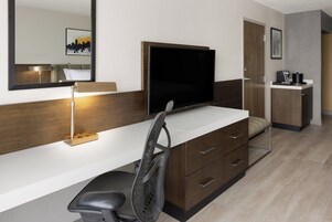 A modern hotel room with a desk, chair, television, and a small kitchenette.
