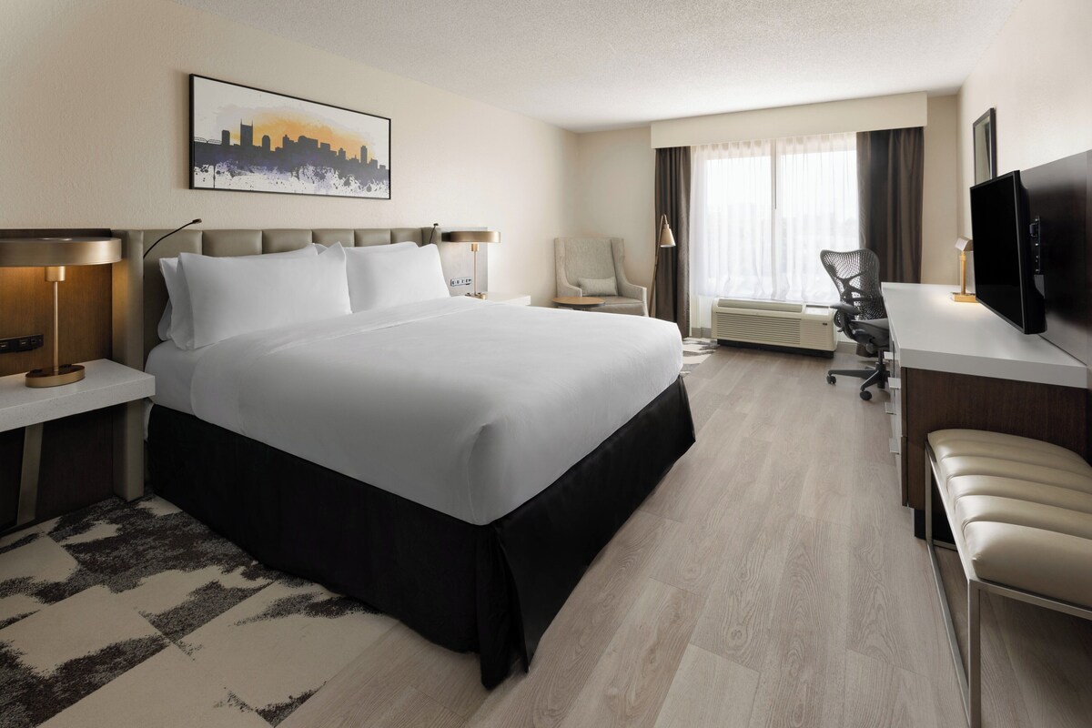A hotel room with a large bed, a desk, a chair, a TV, and a cityscape painting on the wall.