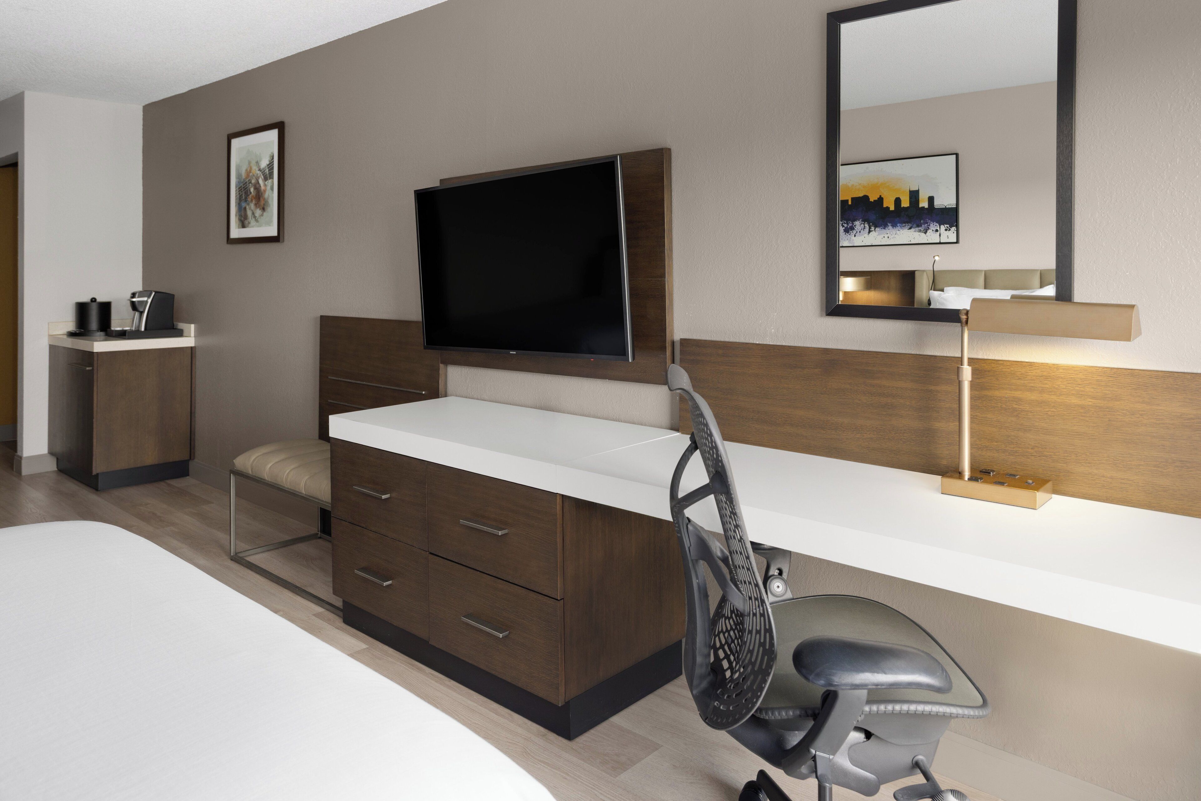 Una habitación de hotel moderna con una cama grande, un escritorio con silla, un televisor y una mesita de noche con lámpara.