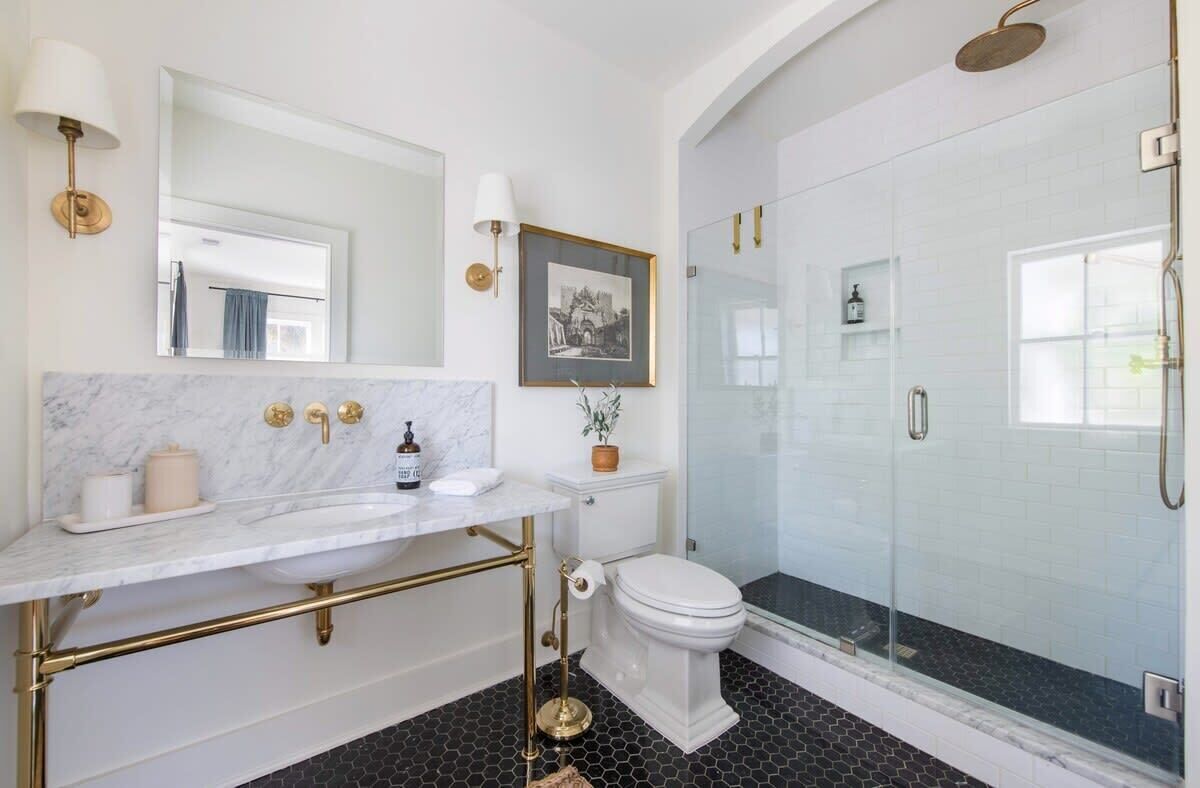 A bathroom with a marble sink, gold fixtures, a toilet, a framed picture, and a glass-enclosed shower.