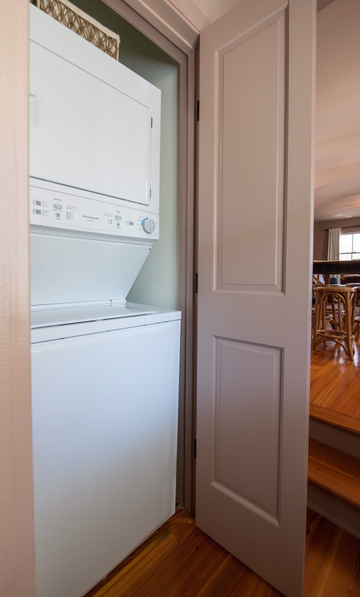 A laundry room with a front-loading washer and dryer, wooden flooring, and a door leading to a dining area with wicker chairs.