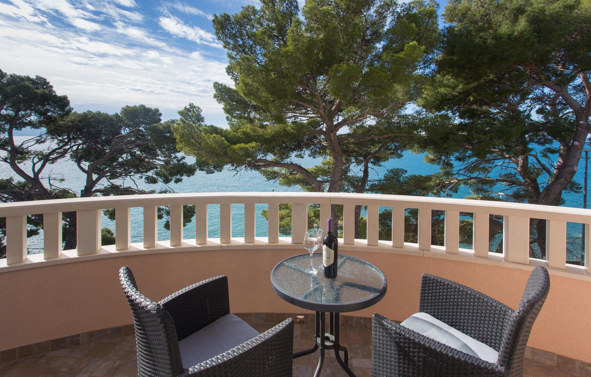 A balcony with wicker chairs and a glass table, overlooking a body of water and trees.
