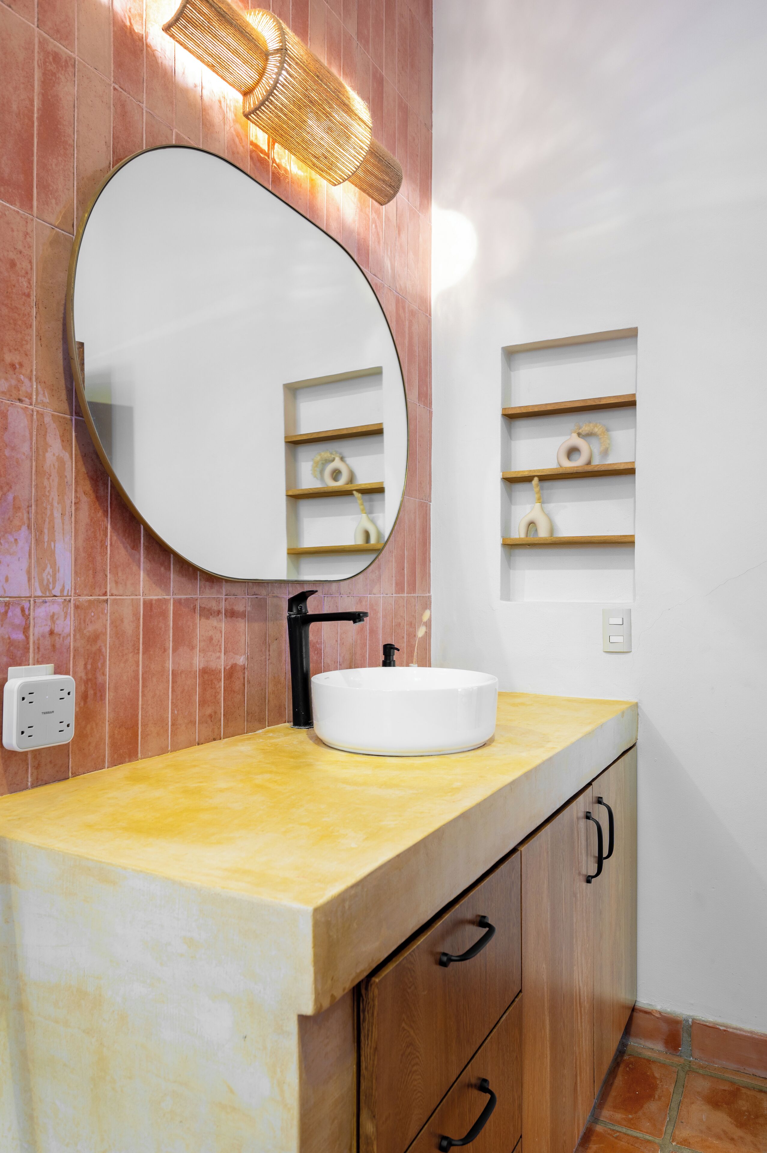 Une salle de bain avec un miroir rond, un lavabo blanc, des meubles en bois et une étagère ornée d’objets décoratifs.