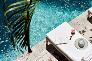 Eine Poolliege mit weißem Liegestuhl, einem Buch, einer Sonnenbrille, einem Getränk und einem Hut.
