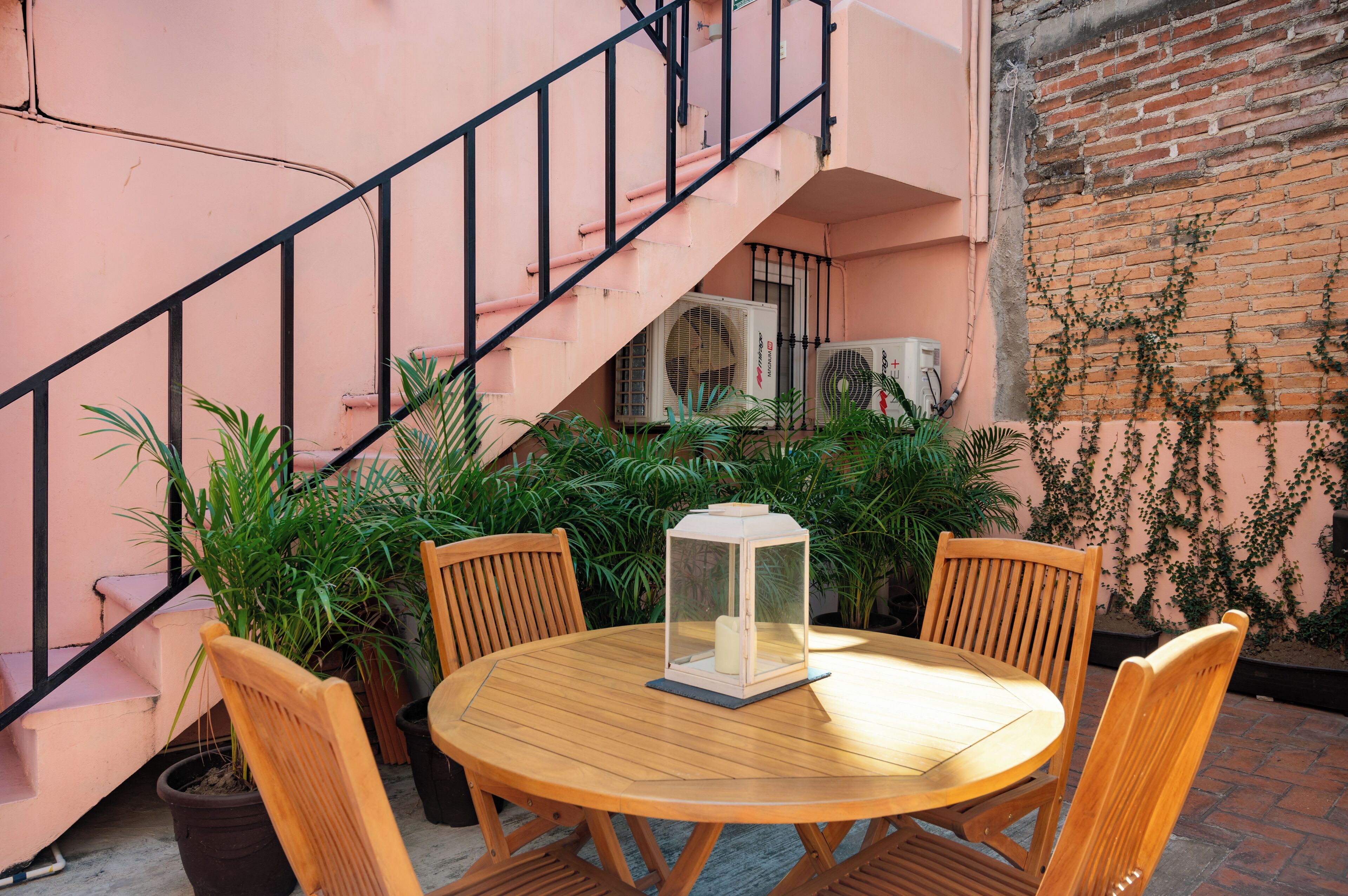 A round wooden table with four chairs, a lantern, and potted plants in an outdoor setting with a pink wall and a brick wall with vines.