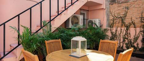 A round wooden table with four chairs, a lantern, and potted plants in an outdoor setting with a pink wall and a brick wall with vines.