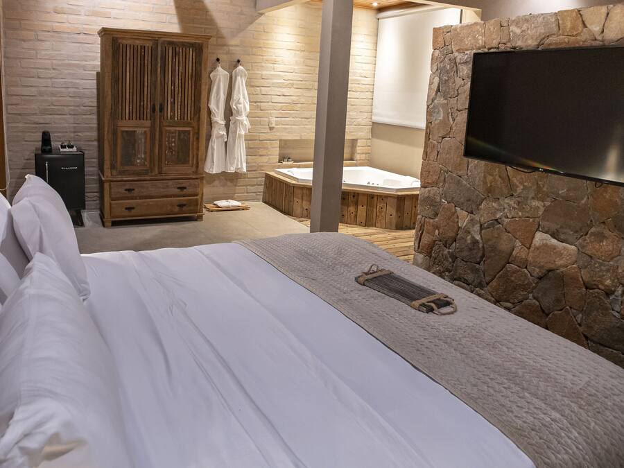 A bedroom with a large bed, a wooden wardrobe, a stone wall, and a flat-screen TV mounted on the wall.