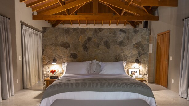 Un dormitorio con pared de piedra, vigas de madera en el techo, cama con ropa blanca, mesita de noche con lámpara y fotografía enmarcada.