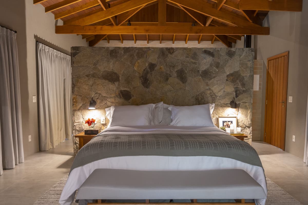 Un dormitorio con pared de piedra, vigas de madera en el techo, cama con ropa blanca, mesita de noche con lámpara y fotografía enmarcada.