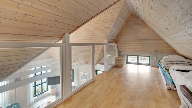 Ein Schlafzimmer im Dachgeschoss mit hohen Decken, Holzbalken, großen Fenstern und einem Bett mit Bettzeug.