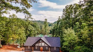 Une maison moderne à deux étages, dotée d’un toit foncé et de grandes fenêtres, entourée d’une forêt dense et accessible par une allée en gravier.
