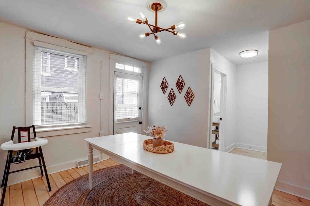 Una habitación amplia con una mesa blanca grande, una silla de fibras naturales y una lámpara en el techo.