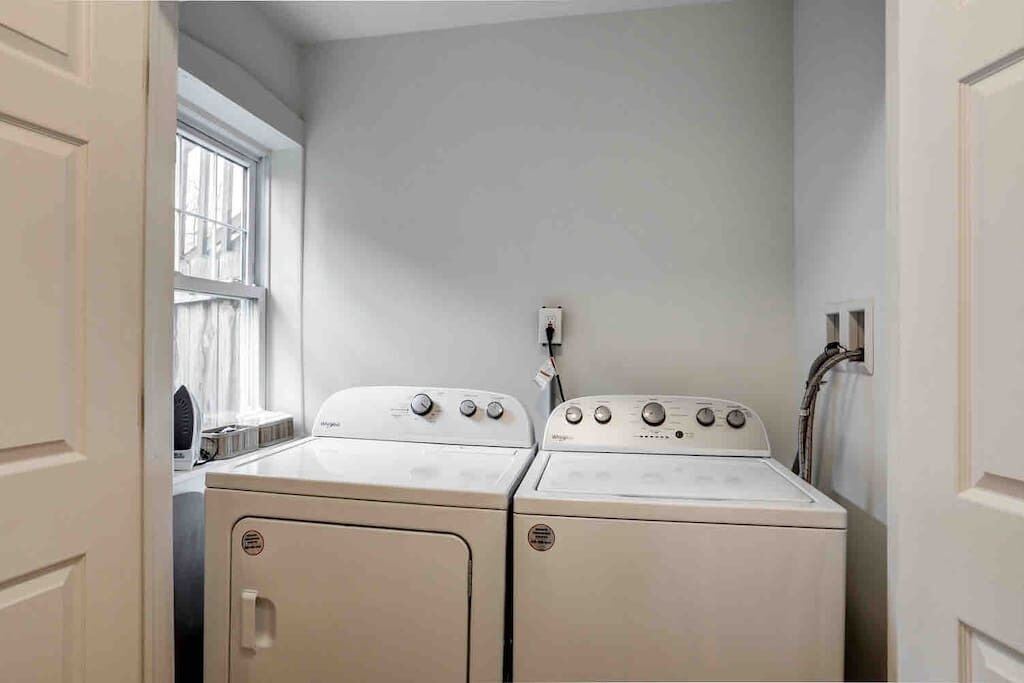A laundry room with two white washing machines and a dryer, a window, and a wall-mounted electrical outlet.