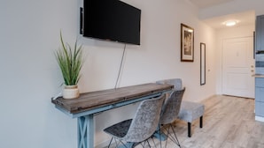 A modern living room with a flat-screen TV mounted on the wall, a wooden dining table, two grey upholstered chairs, a potted plant, and a framed picture on the wall.