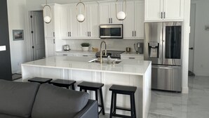 A modern kitchen with white cabinets, stainless steel appliances, and a central island with bar stools. - New 4bd/3ba w/ private pool, fenced yard, outdoor kitchen, close to everything! (Cape Coral)