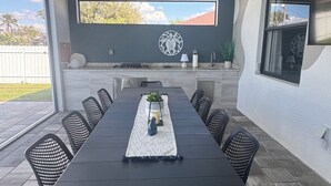 An outdoor patio with a long dining table, chairs, and a ceiling fan. - New 4bd/3ba w/ private pool, fenced yard, outdoor kitchen, close to everything! (Cape Coral)
