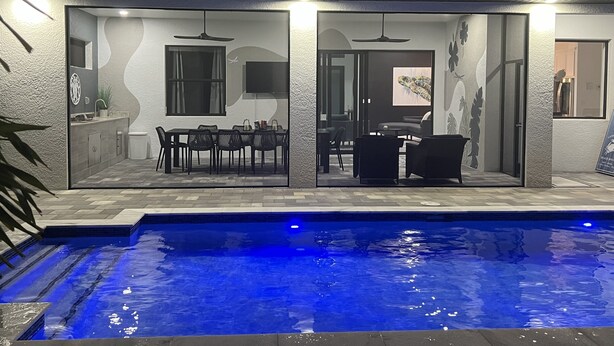 A modern outdoor pool area with a dining setup and a kitchen visible through glass doors. - New 4bd/3ba w/ private pool, fenced yard, outdoor kitchen, close to everything! (Cape Coral)