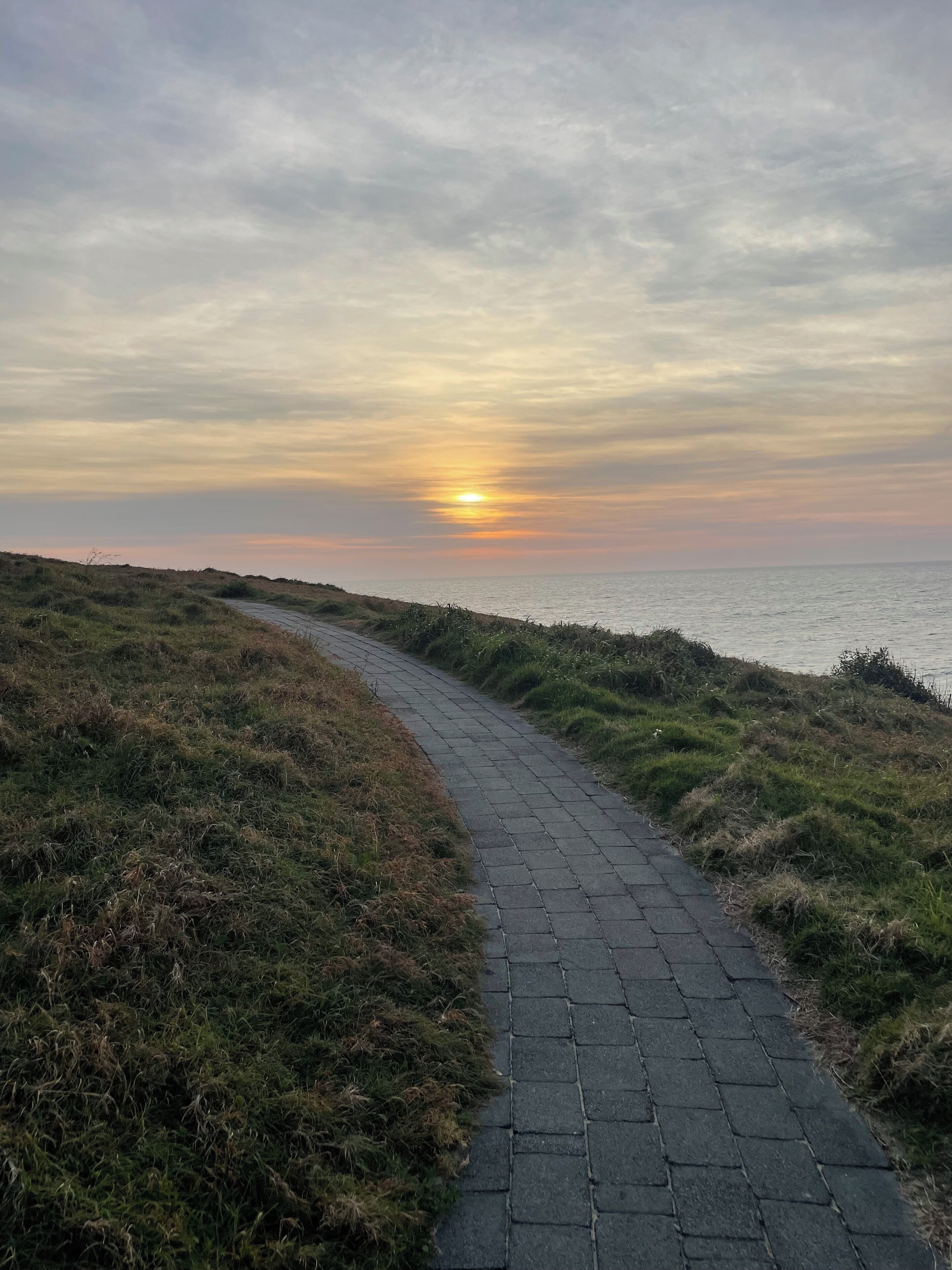 Un sendero pavimentado serpentea a lo largo de una ladera cubierta de hierba, con vistas a la puesta de sol sobre el agua.