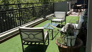 Ein Balkon mit künstlichem Rasen, einem Tisch aus Metall, zwei weißen Sitzgelegenheiten mit Polstern und einem Blumentopf.