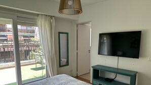 Ein Schlafzimmer mit einem Bett, einem Fernseher auf einem türkisblauen Schrank, einer Schiebetür und Blick ins Freie.