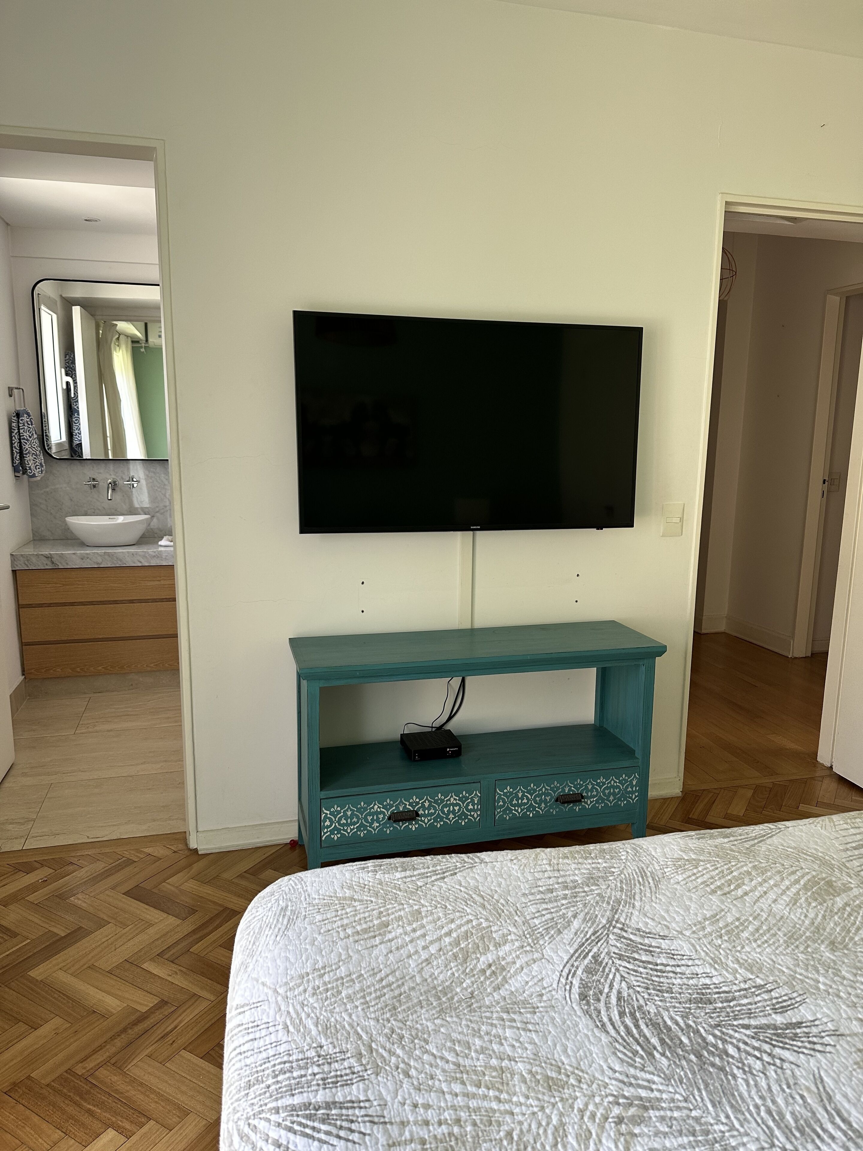 A bedroom with a bed, a TV mounted on a teal console, a bathroom visible through an open door, and a wooden floor.