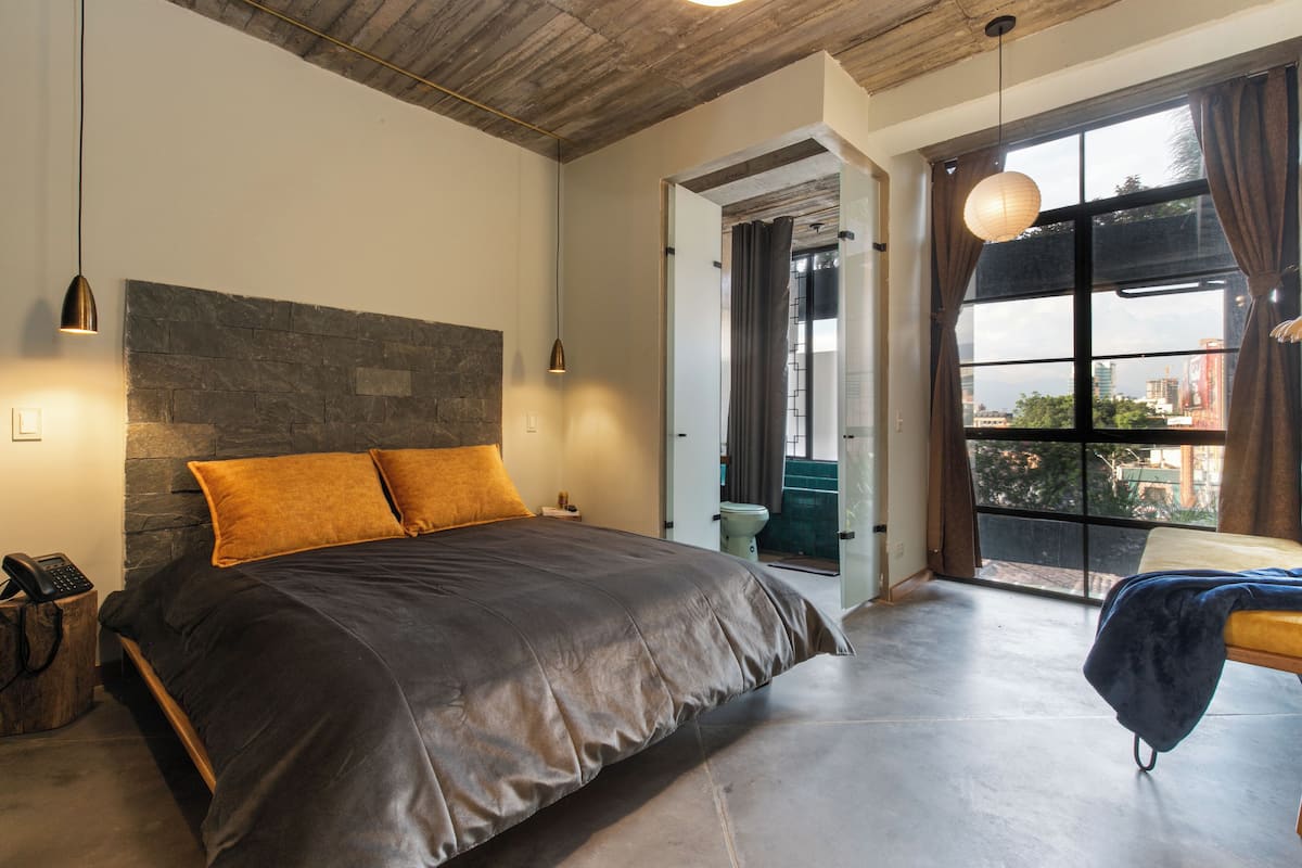 A modern bedroom with a large bed, stone wall, and a view of the city through a large window.