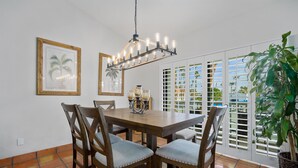 Un comedor con una mesa y sillas de madera, una lámpara de araña, cuadros enmarcados y vistas a través de una ventana con contraventanas.