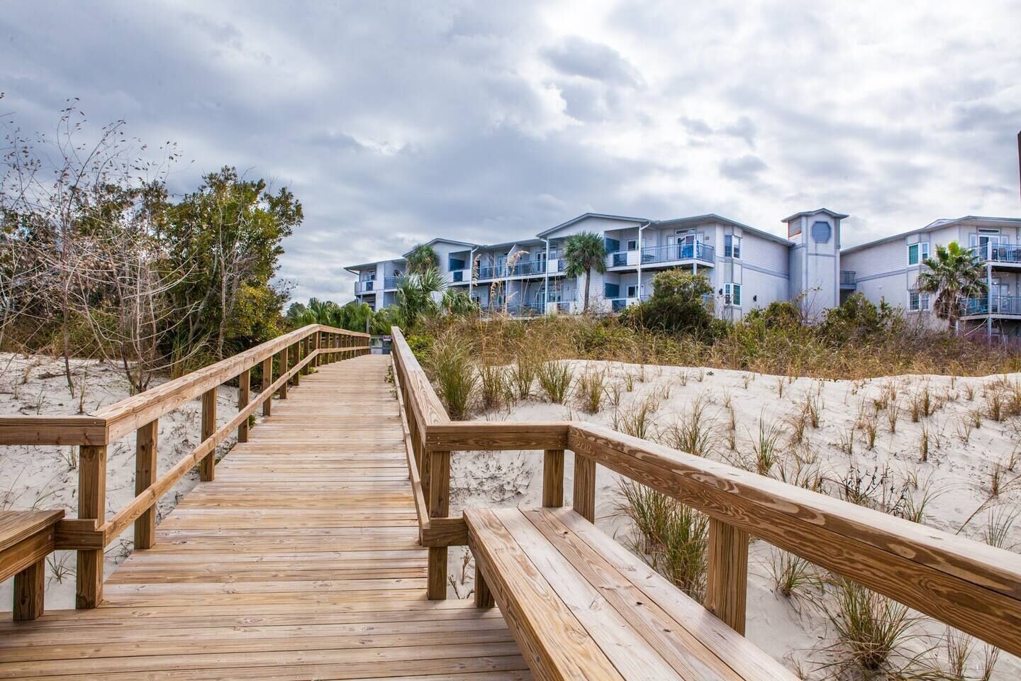 A wooden boardwalk leading to a beachfront hotel with multiple balconies and a clock tower.