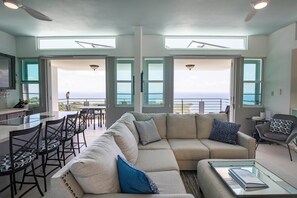 A modern living room with a large sectional sofa, a dining area with bar stools, and a kitchen area. The room has large windows offering a view of the ocean.
