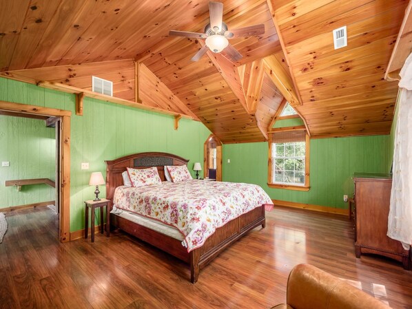 Une chambre spacieuse avec un plafond en bois, un grand lit, une table de chevet, une commode et une fenêtre donnant sur les arbres.