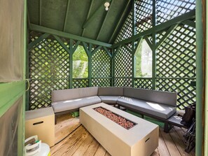 Un espace extérieur aménagé, partiellement abrité, avec un foyer intégré, un plancher en bois et des murs recouverts d’un treillis végétal.