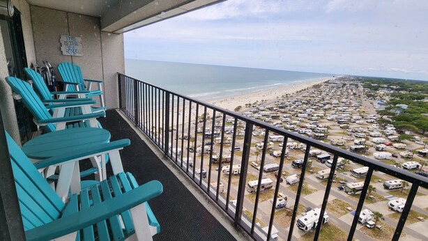 A balcony with turquoise Adirondack chairs, a view of a beach, and a parking lot with RVs.